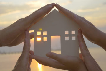 Group of human palms on all sides of a cut out house diagram with setting sun coming through window over water